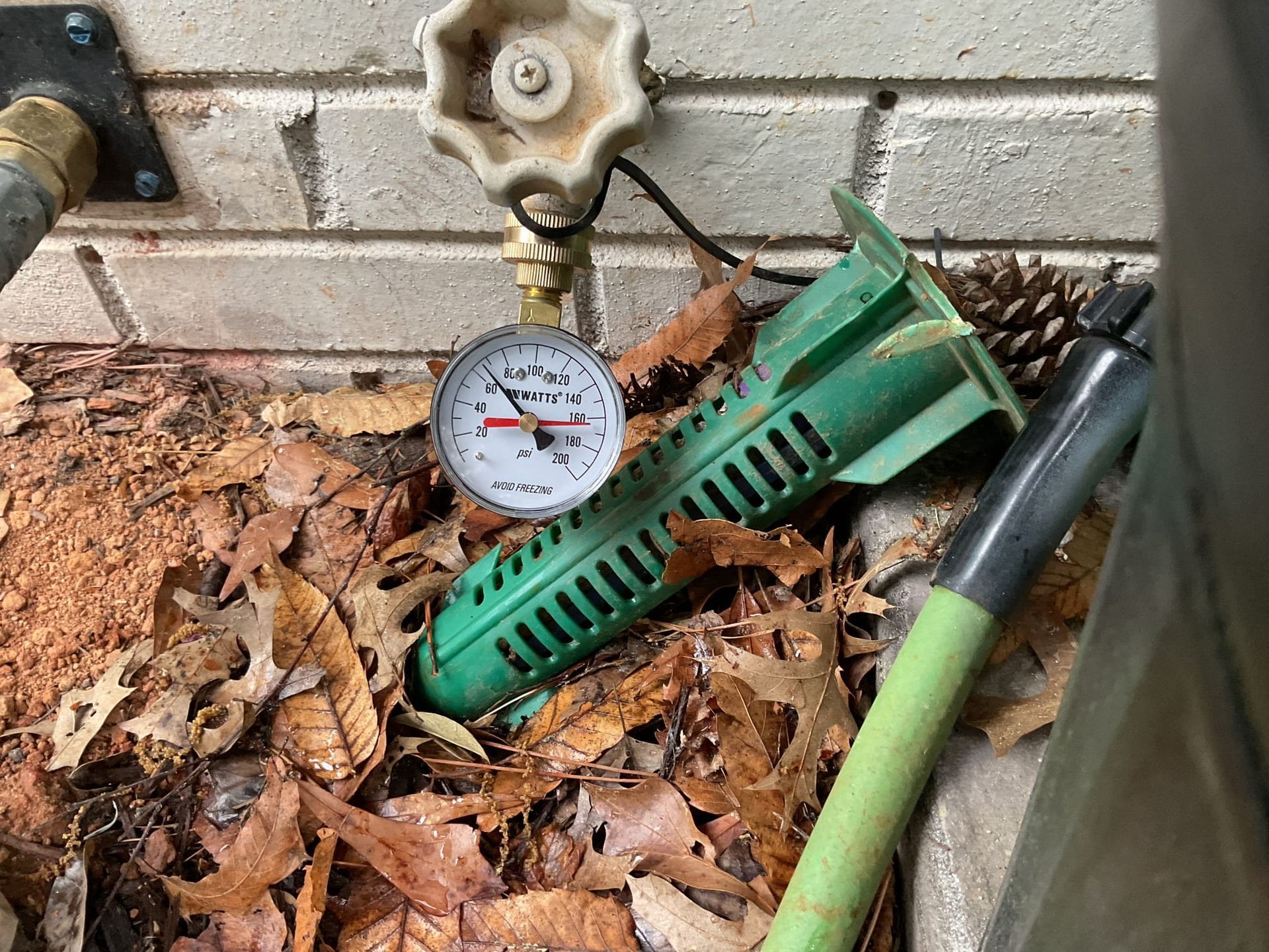 Water pressure gauge near a spigot and green sprinkler, surrounded by leaves and brick.
