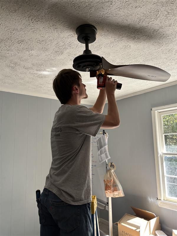 Person installing a ceiling fan, using a tool. He is on a ladder in a room with blue walls and a window.