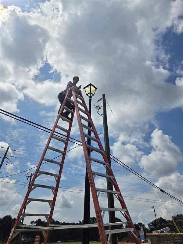 Person on a ladder, working on a lamppost against a cloudy sky.