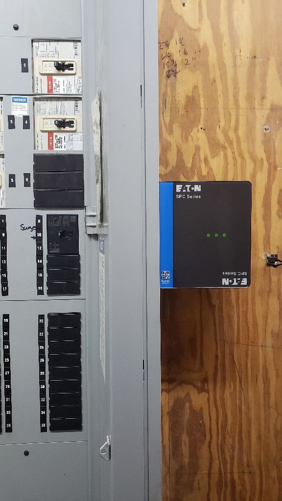 Electrical panel and small black and blue device mounted on wood panel.