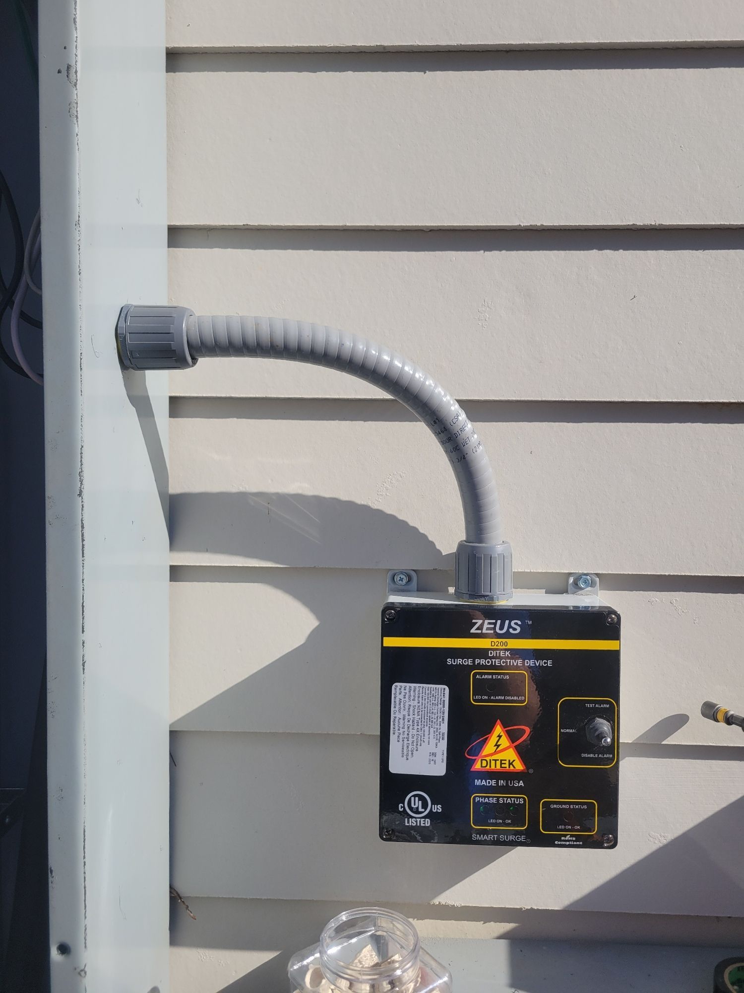 Electrical control box mounted on a light beige siding, gray conduit attached, with a warning label.