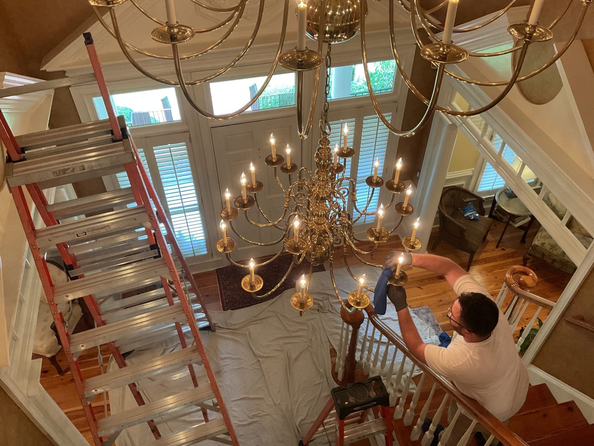 Person cleaning a large chandelier, standing on a ladder. White cloth covers floor.