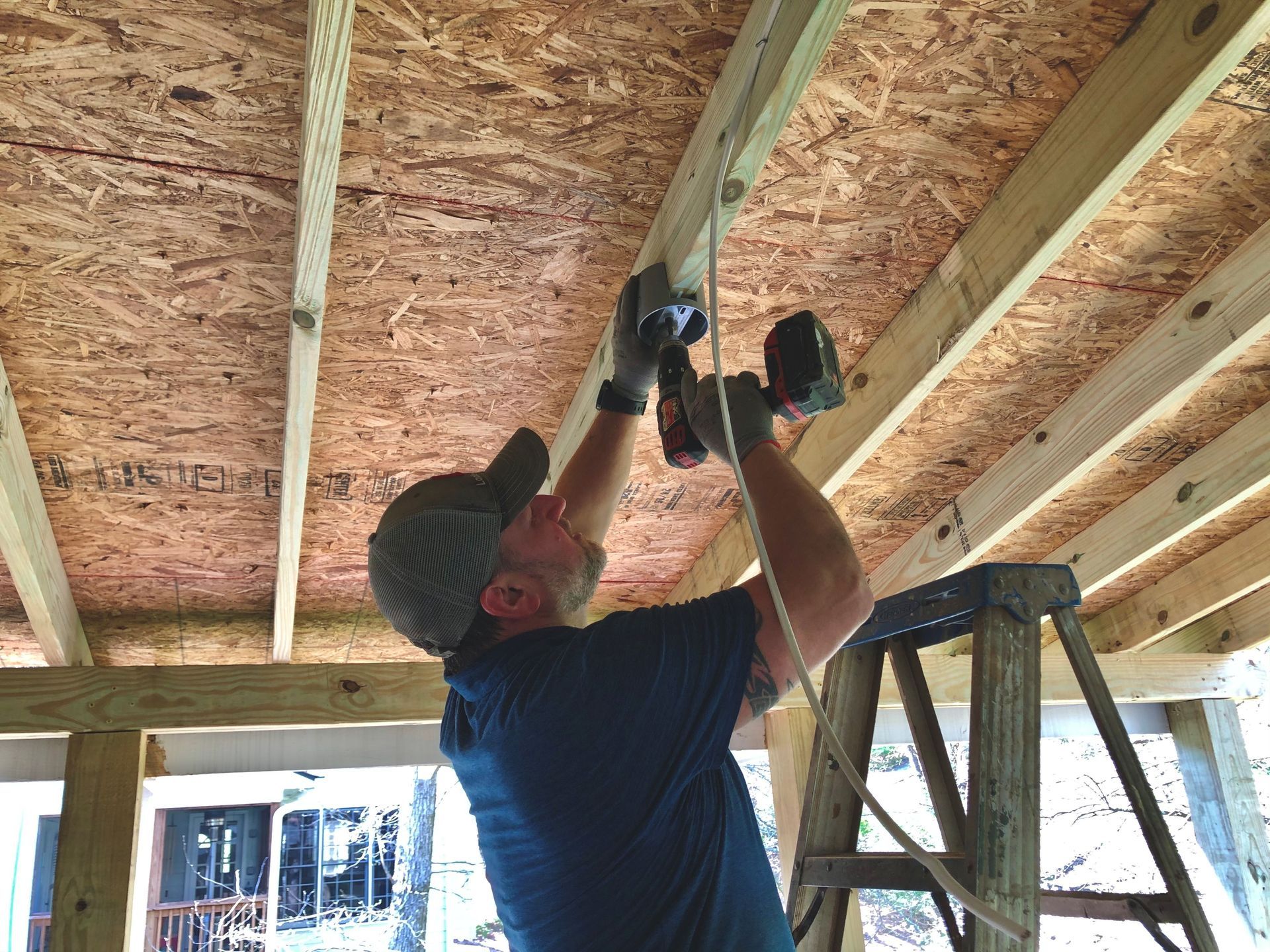 Person on a ladder using a power drill to install something on a wooden ceiling.