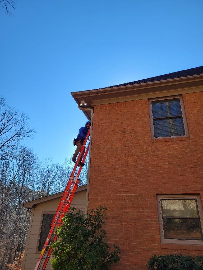 Person on a red ladder cleaning gutters on a two-story brick building. Clear blue sky in background.
