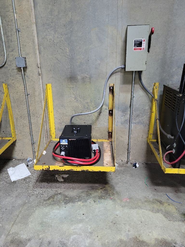 A black box, electrical panel, and wiring on a yellow shelf attached to a concrete wall.