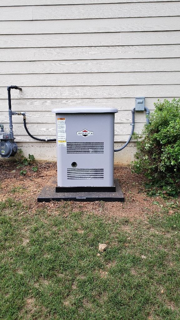 Outdoor generator next to a house with gas and electrical connections, surrounded by grass and bushes.
