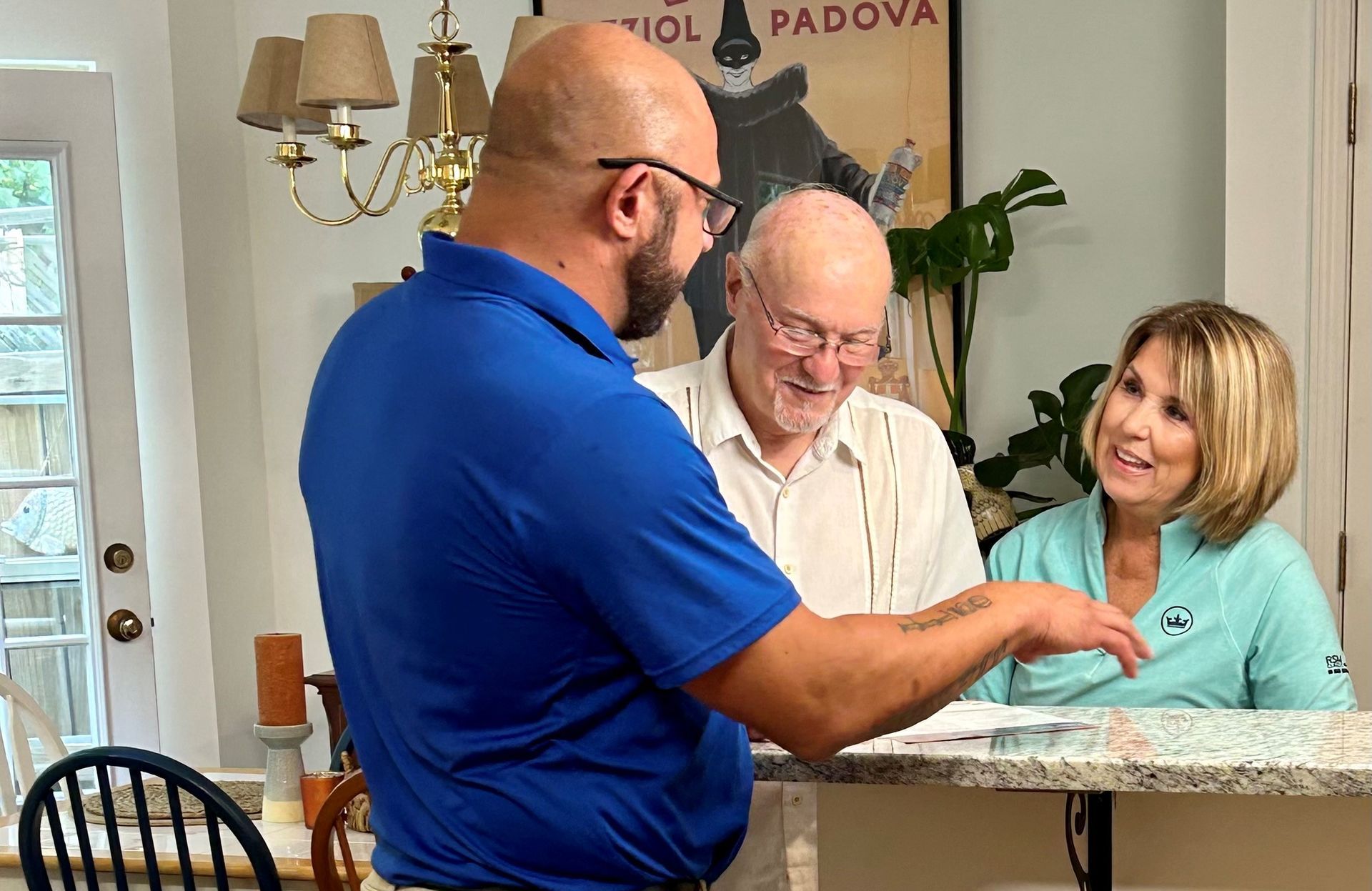 Three people at a counter; man gestures, older man smiles, woman smiles. Interior, poster & plant visible.