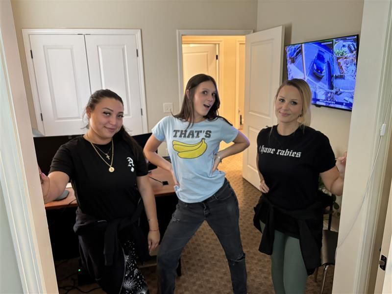 Three women pose in a room. One wears a 