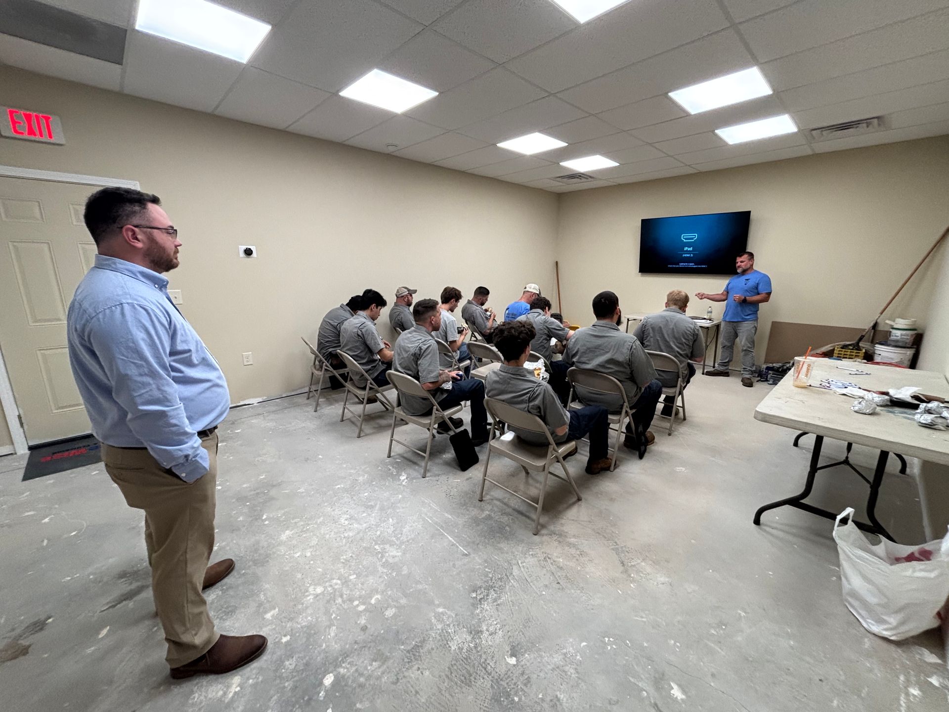 Group of people in a room attending a presentation, one man stands, others seated, a presenter gestures at a screen.