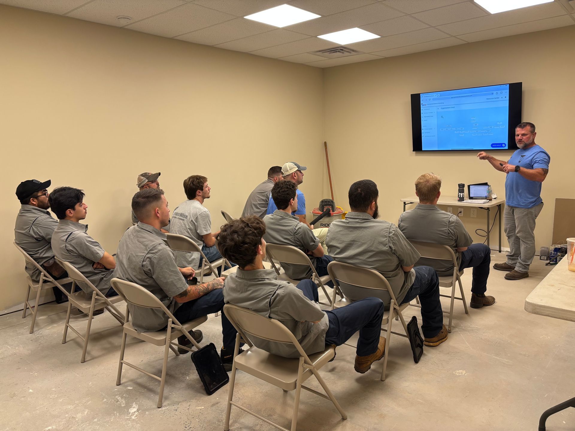 A man presents to a group of people seated in a classroom; a screen is behind him.
