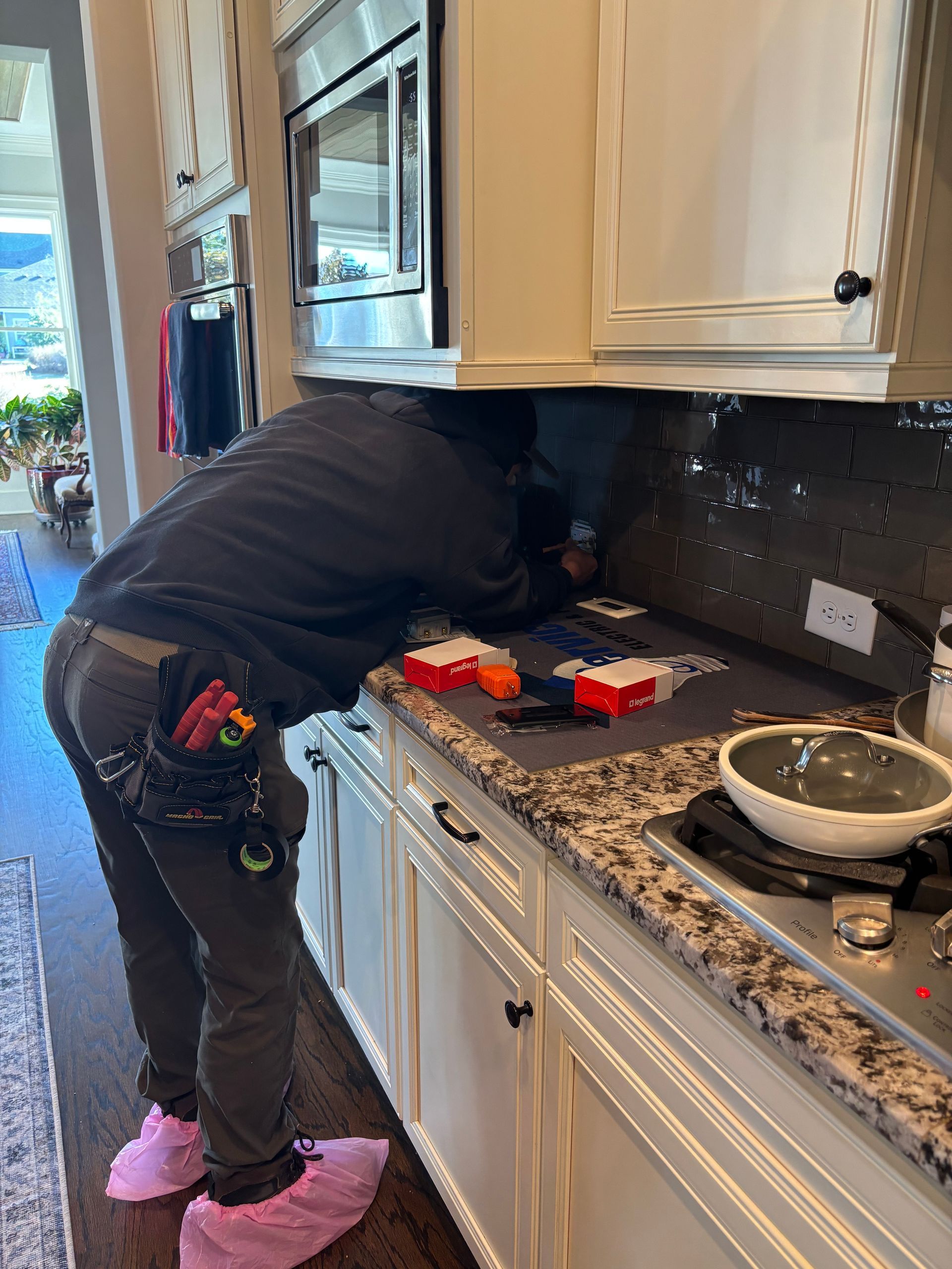 Person working under a kitchen cabinet, wearing pink shoe covers, with tools in a pouch.