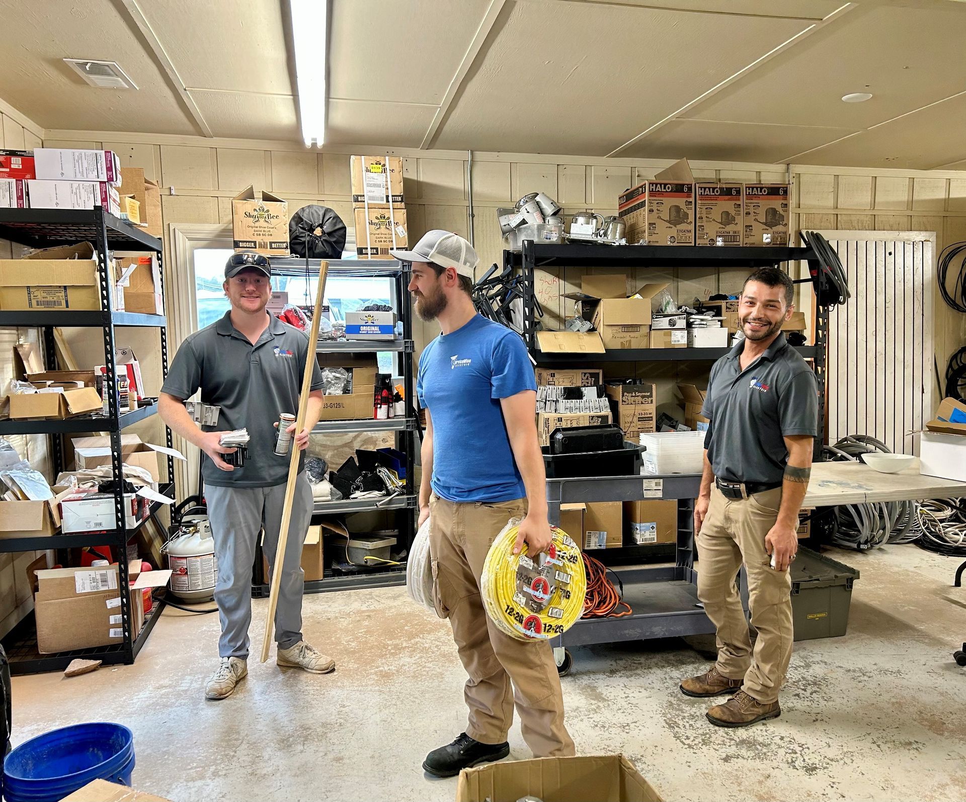 Three men in a cluttered warehouse setting; one holds cable, another a pole, and the third a canister.