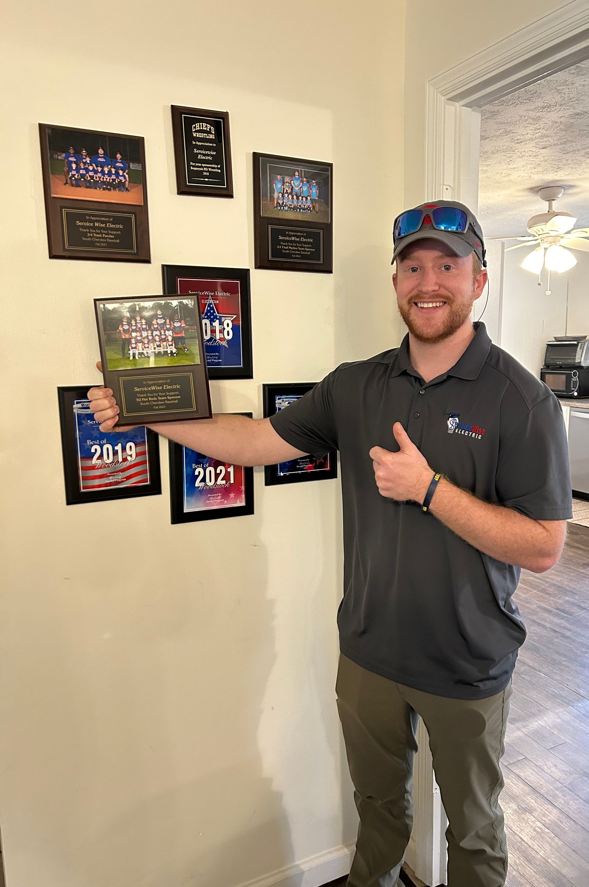 Man holding award, giving thumbs up, in front of wall with other plaques. Room with door and overhead light.