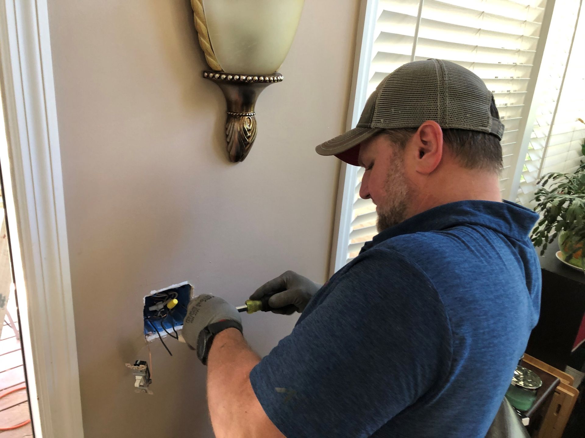 Electrician installing a wall outlet. He wears a hat and gloves, working on wires with tools on a beige wall.