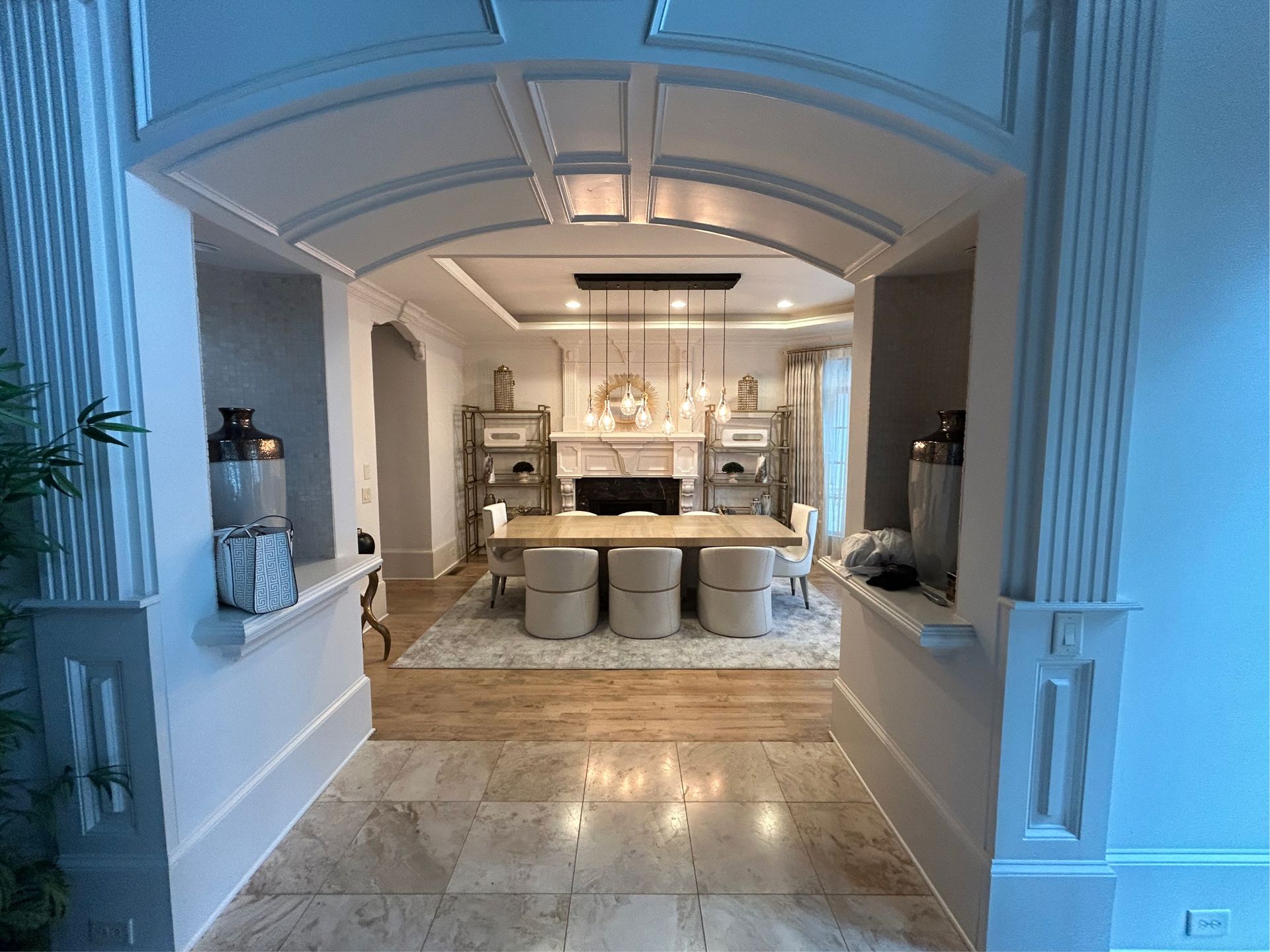 An entryway with a dining room in the background, cream chairs, wood table, arched ceiling, and stone floor.