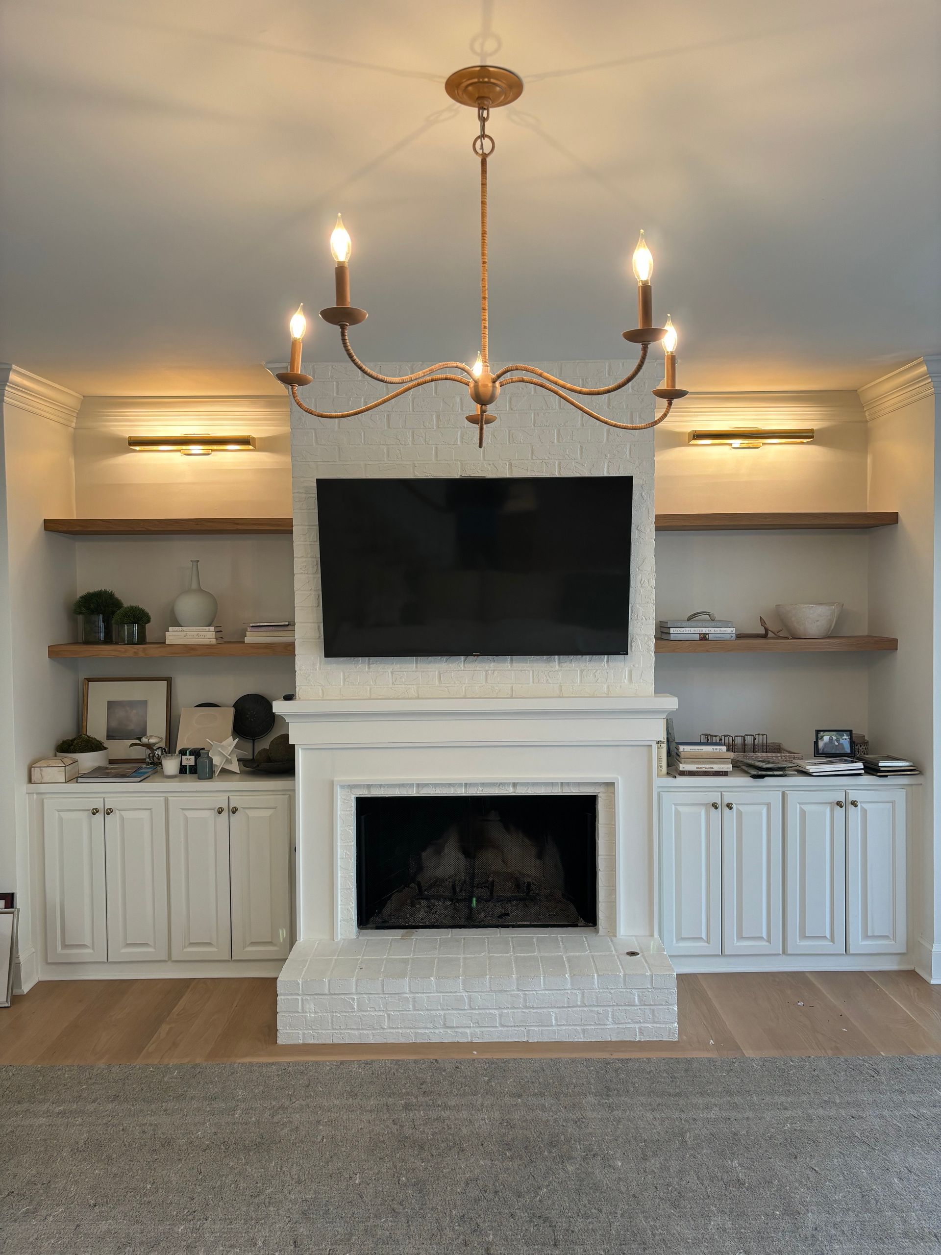Living room with fireplace, built-in shelves, TV, and gold chandelier. White brick fireplace, light wood shelves, light grey carpet.