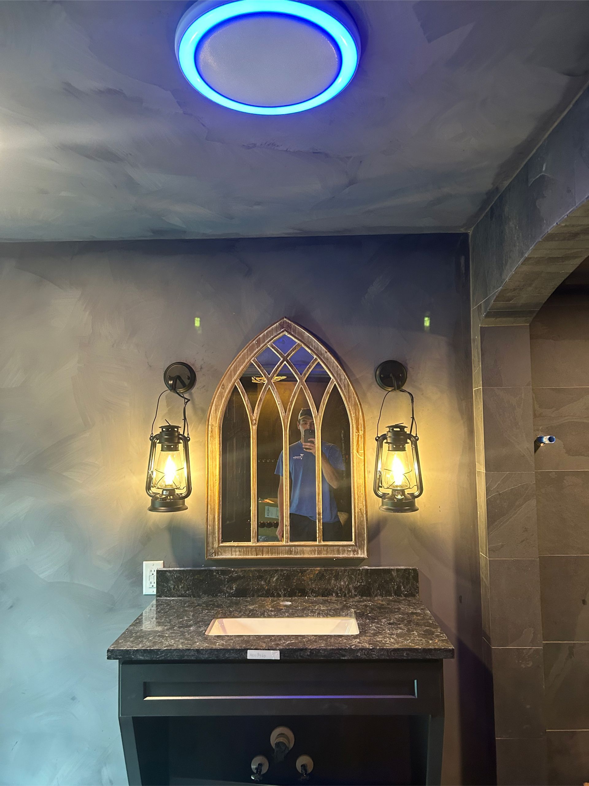Vanity with ornate mirror, lantern sconces, and blue-lit ceiling fixture against a dark, textured wall.