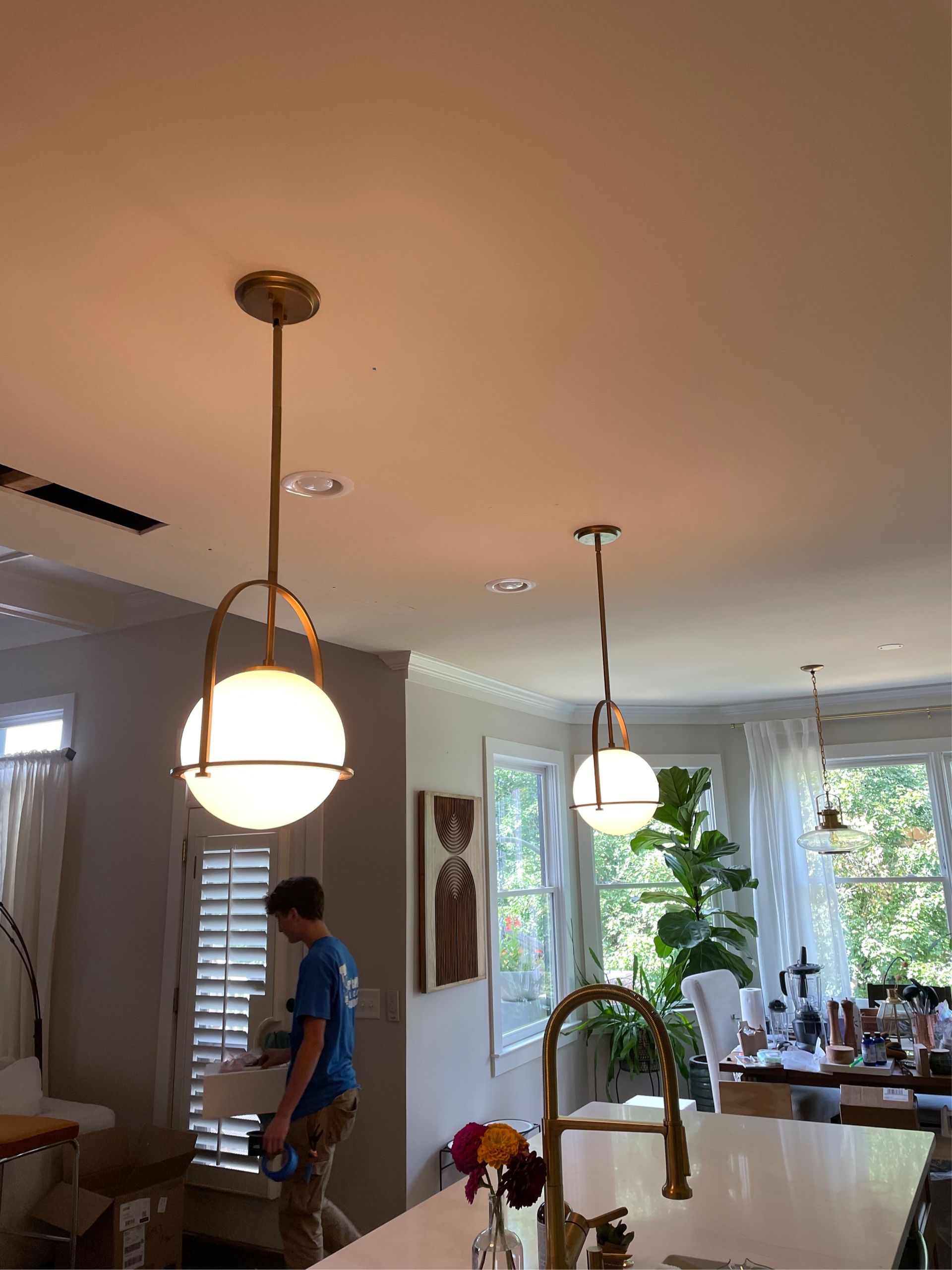 Two gold pendant lights with white globes hang over a kitchen island. A person walks by a window.