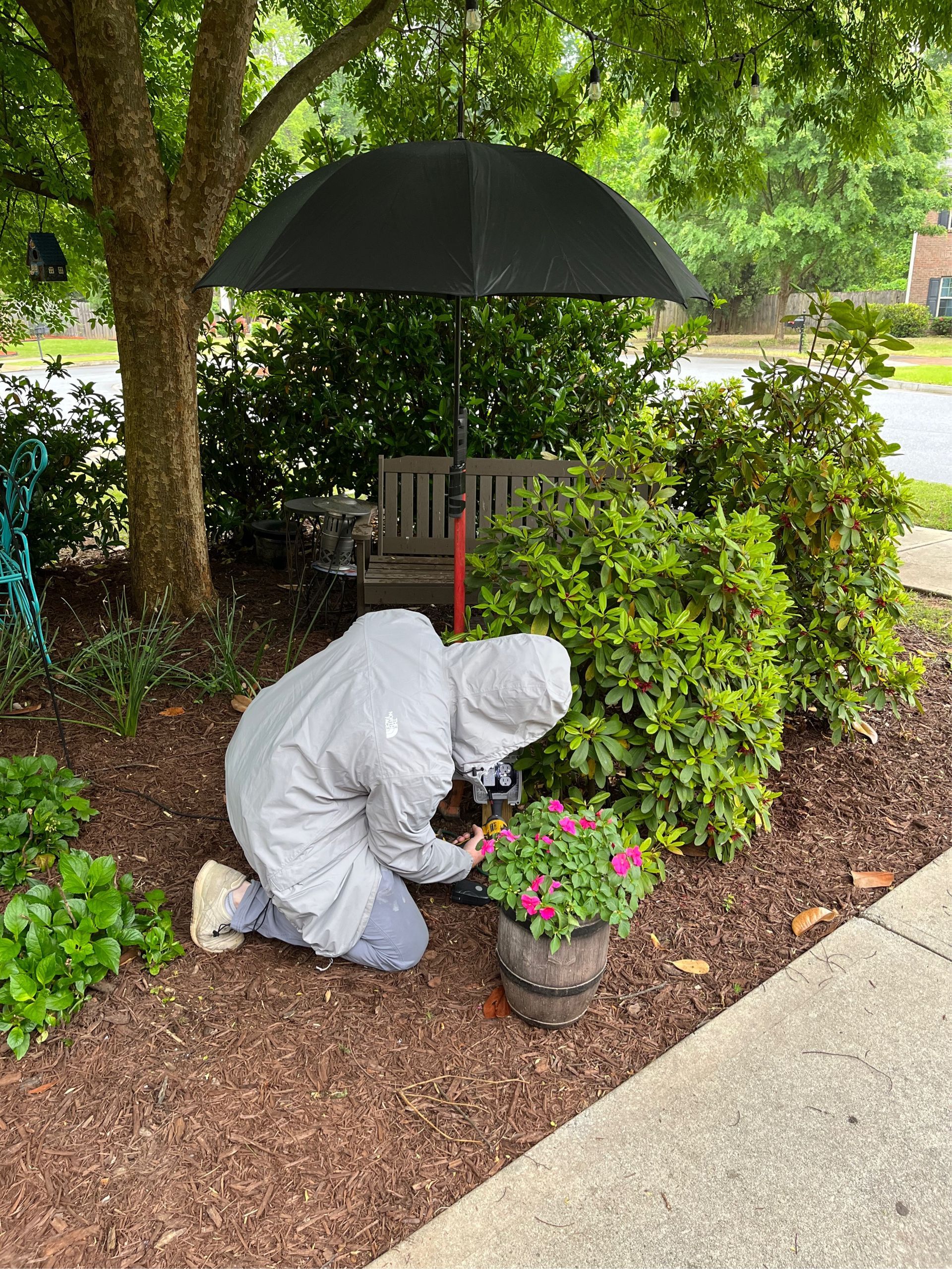 Person in a raincoat kneels to tend flowers under an umbrella in a garden.