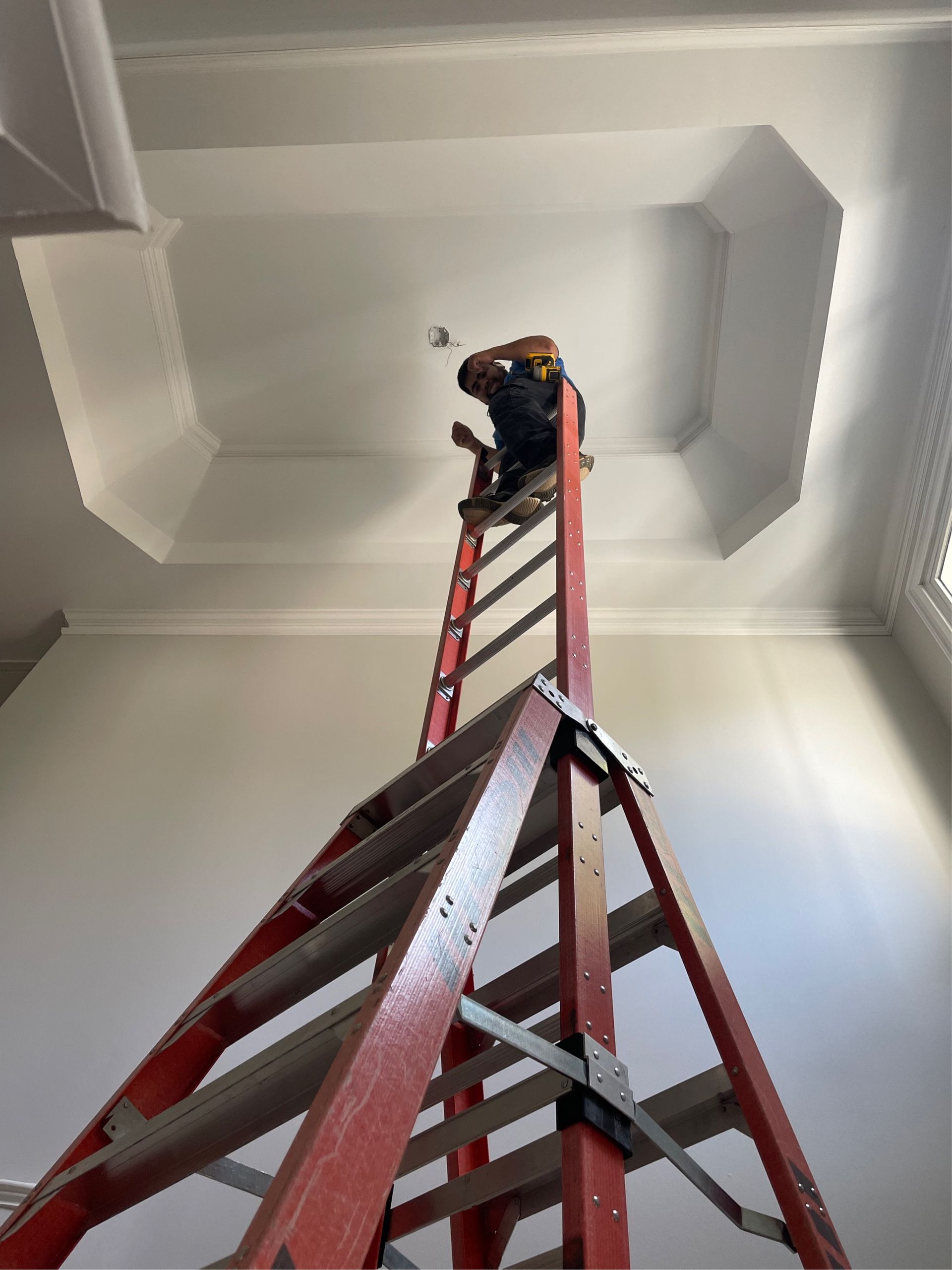 Person on a red ladder working on a ceiling fixture in a room with white walls and ornate trim.