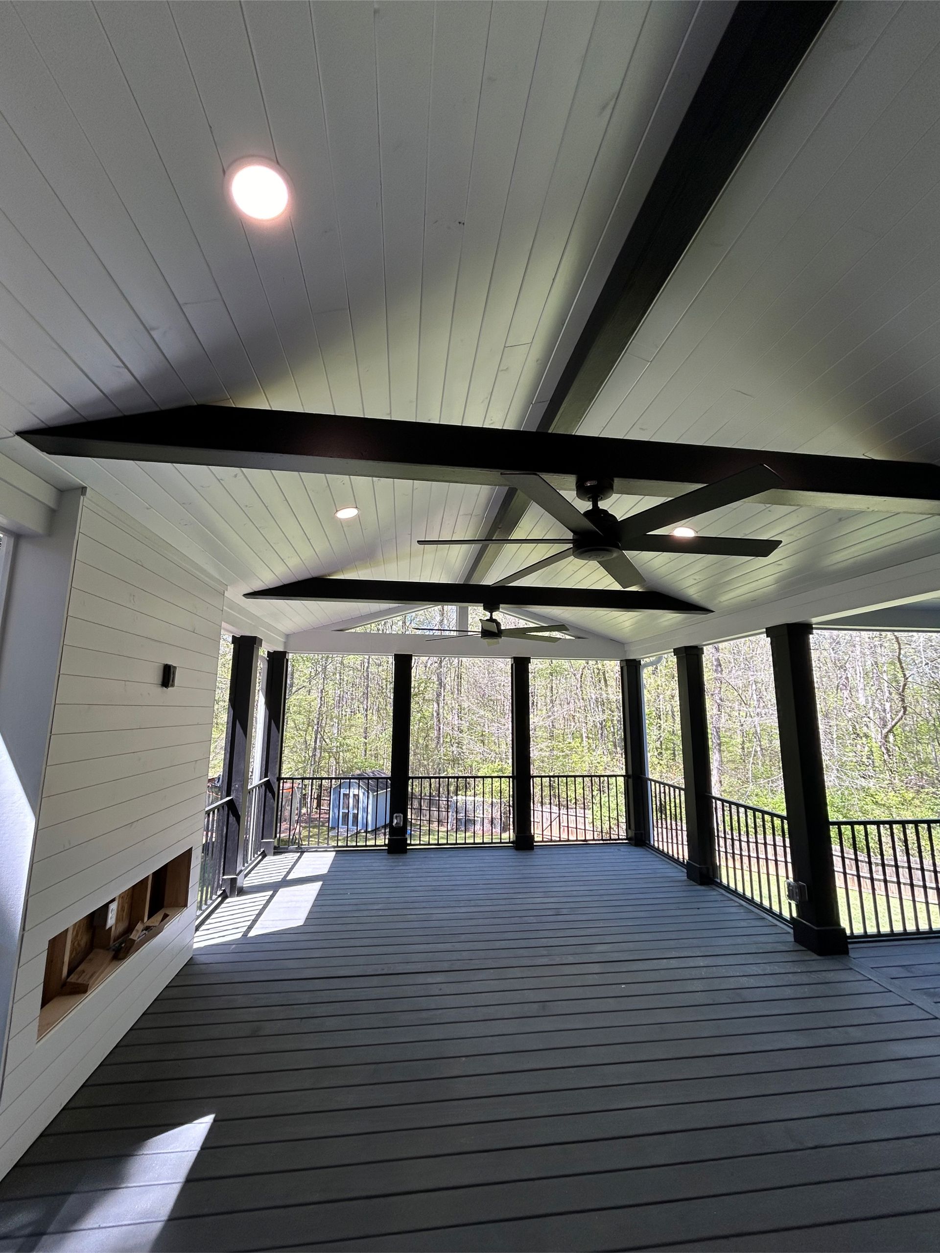 Covered porch with dark beams, ceiling fan, and black railing overlooking a wooded area.