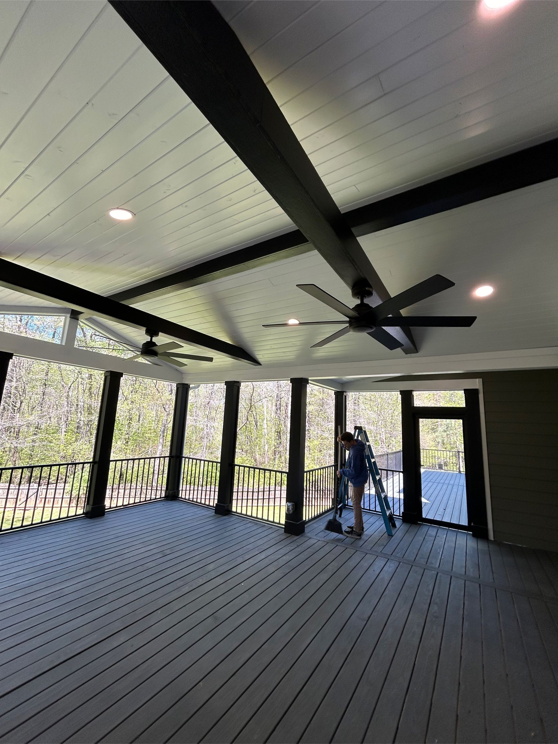 Covered outdoor deck with grey flooring, white ceiling with black beams, and a person working near a doorway.