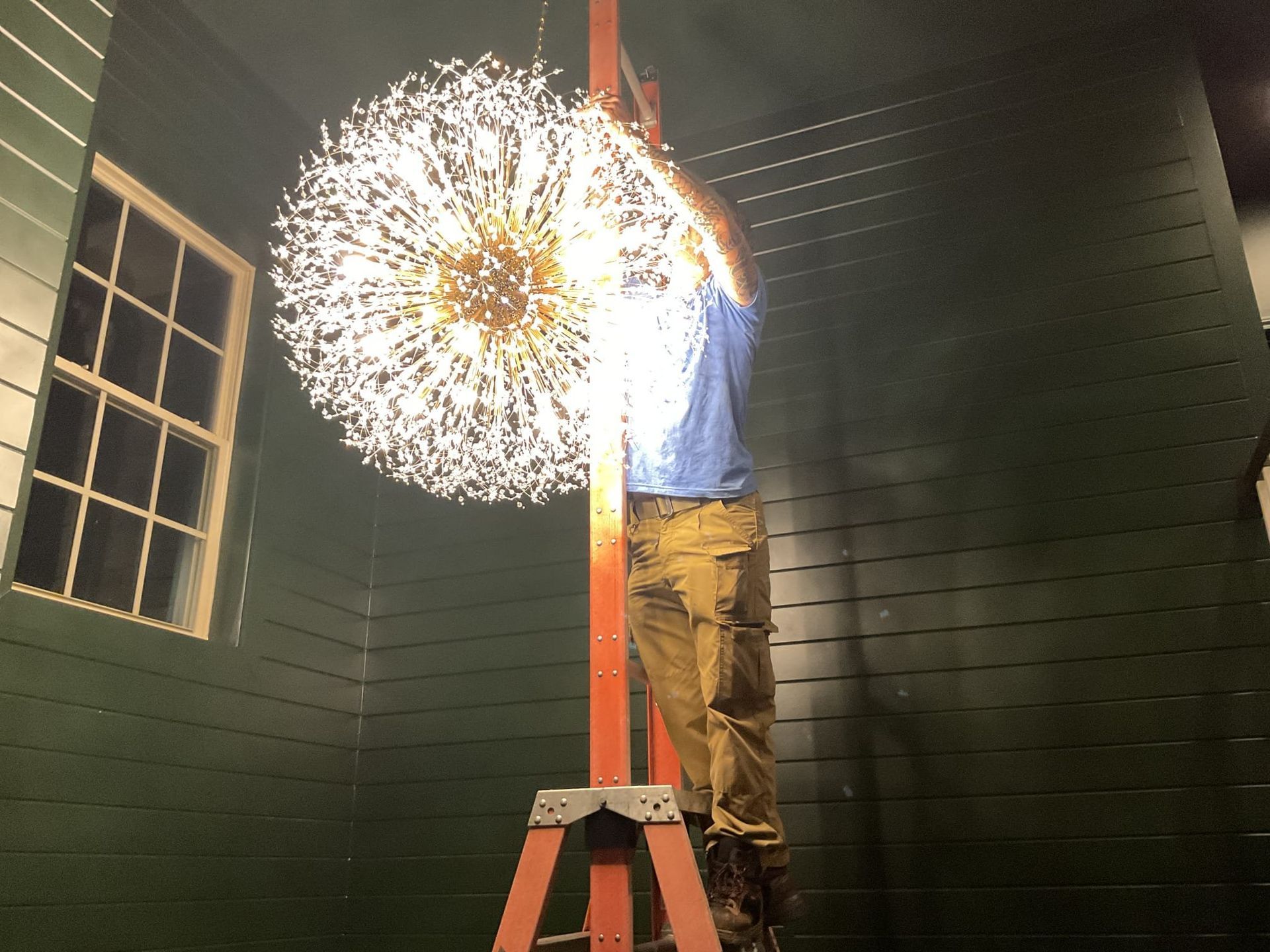 Person on a ladder installing a large, decorative, illuminated sphere in a room with a window.