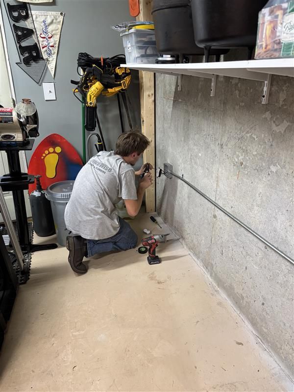 Person kneels, using a drill on a metal pipe attached to a concrete wall in a garage.