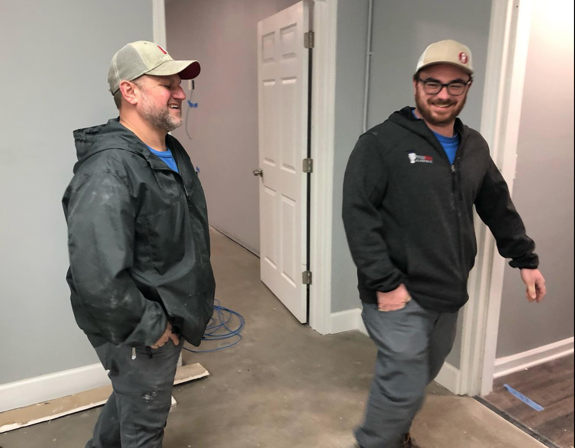 Two men in a room, one smiles, both wear caps and jackets. Gray walls, open door.