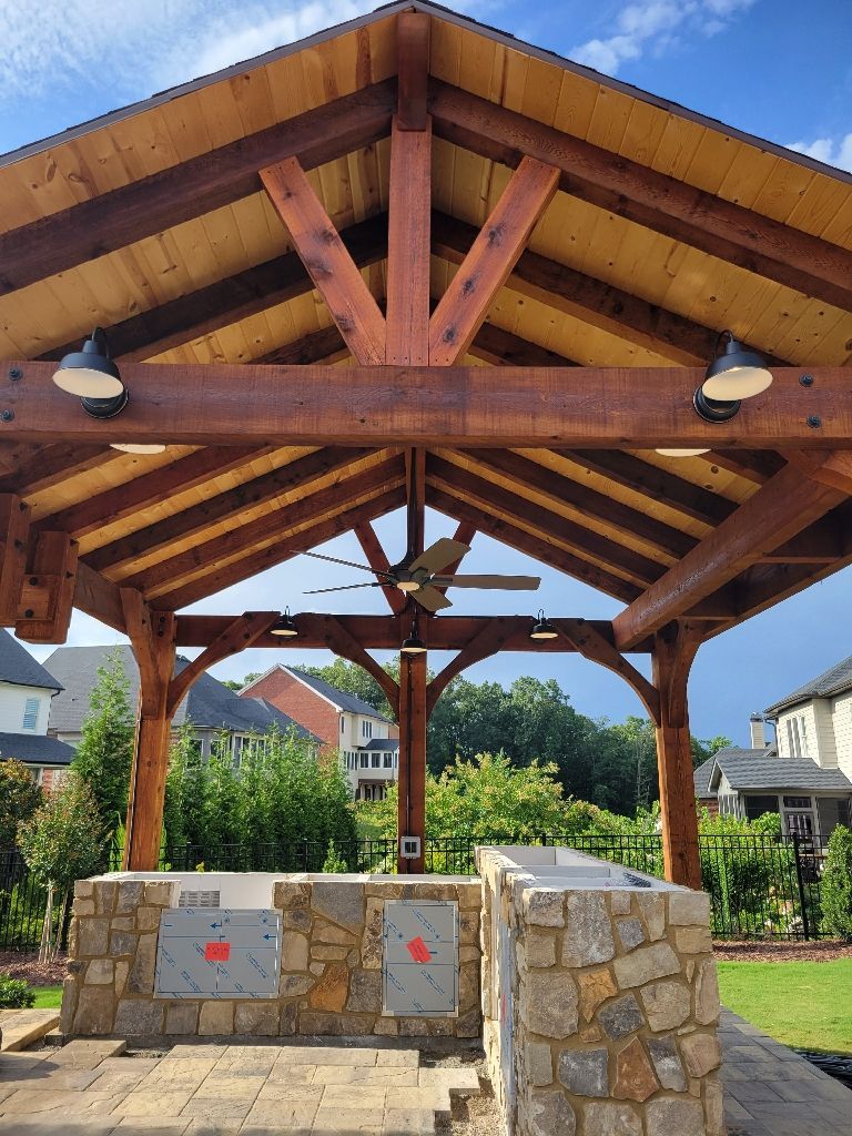 Wooden pergola with stone grill station, fan, and lights. Houses and trees in the background.