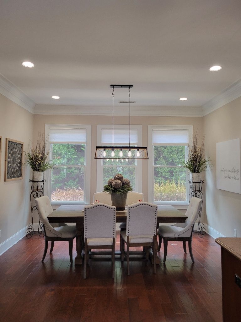 Dining room with a wooden table, six upholstered chairs, and a chandelier. Windows with greenery visible outside.