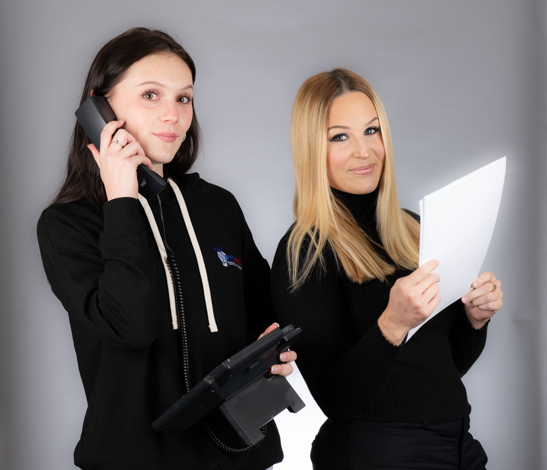 Two women, one on phone, the other holding papers, both wearing black clothing. Studio setting.