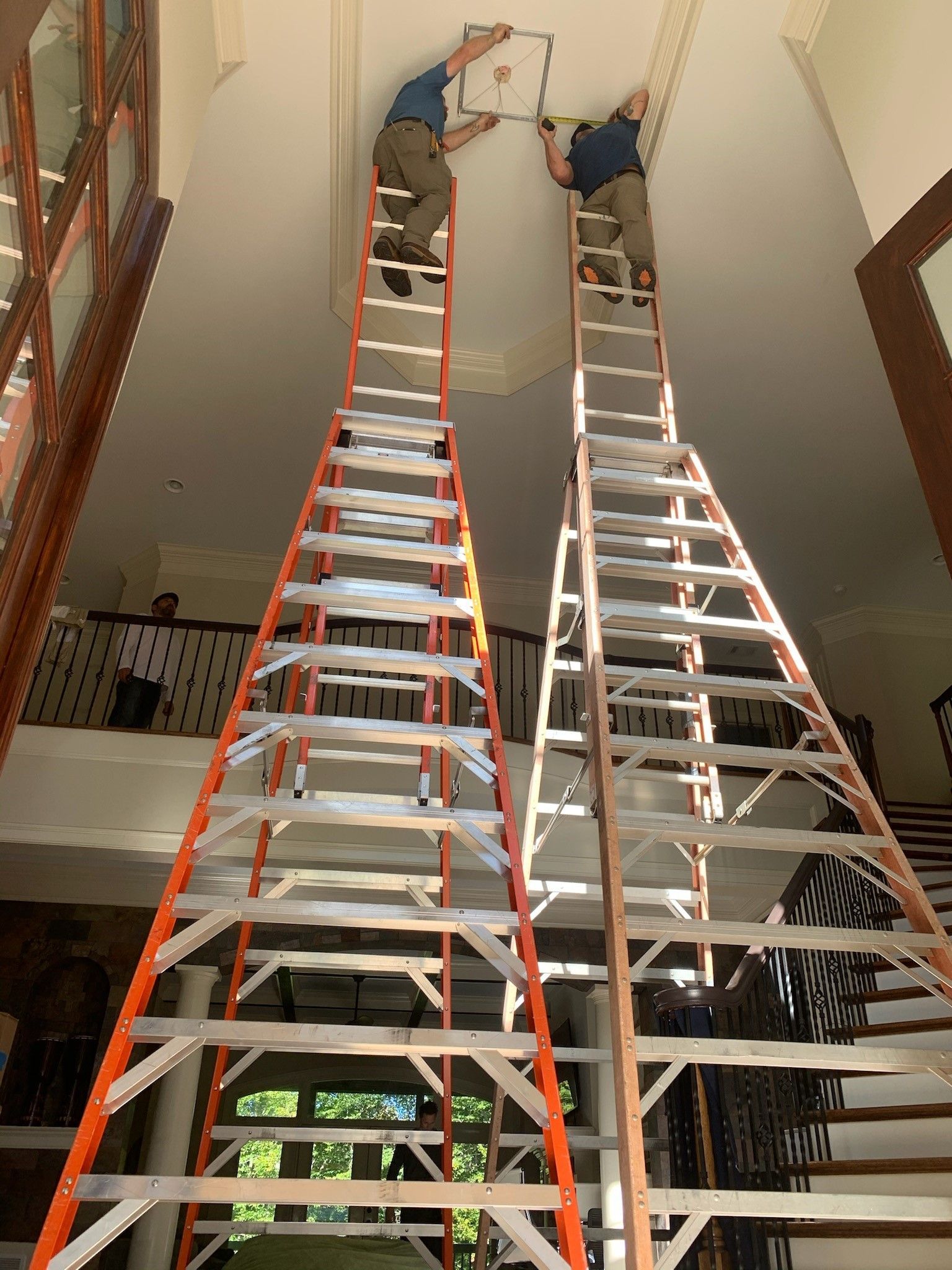 Two workers on tall ladders install something on a high ceiling. Interior setting, well lit.