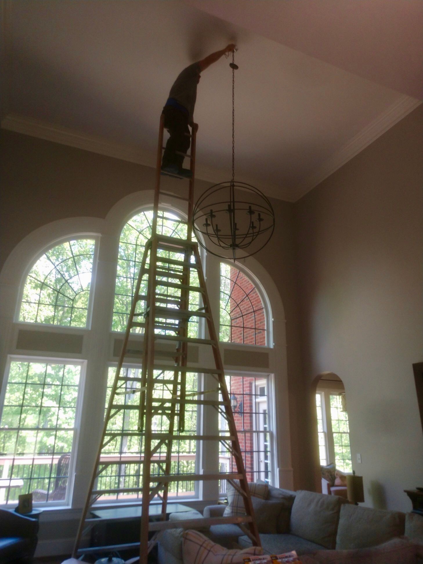 Person on tall ladder adjusting a chandelier in a room with large windows.