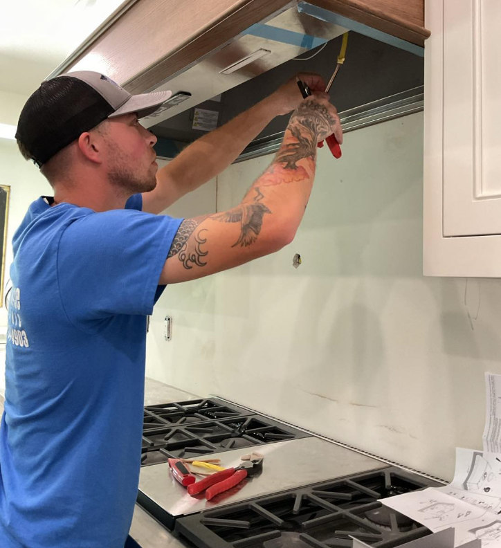 Person in a blue shirt installing a range hood above a stovetop in a kitchen, using a screwdriver.