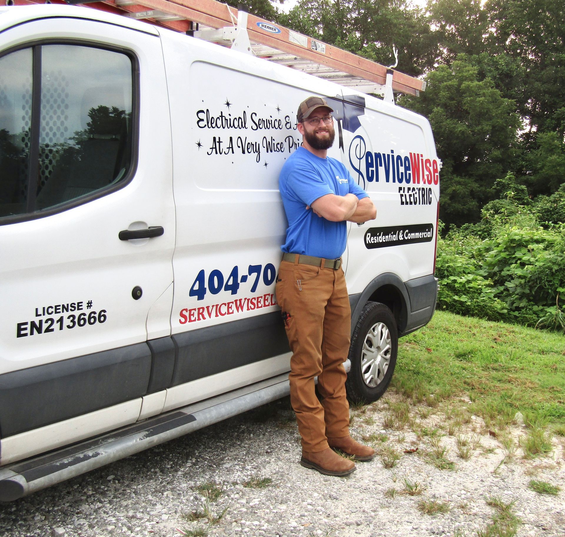 Man in work clothes leans against a white van, arms crossed. 