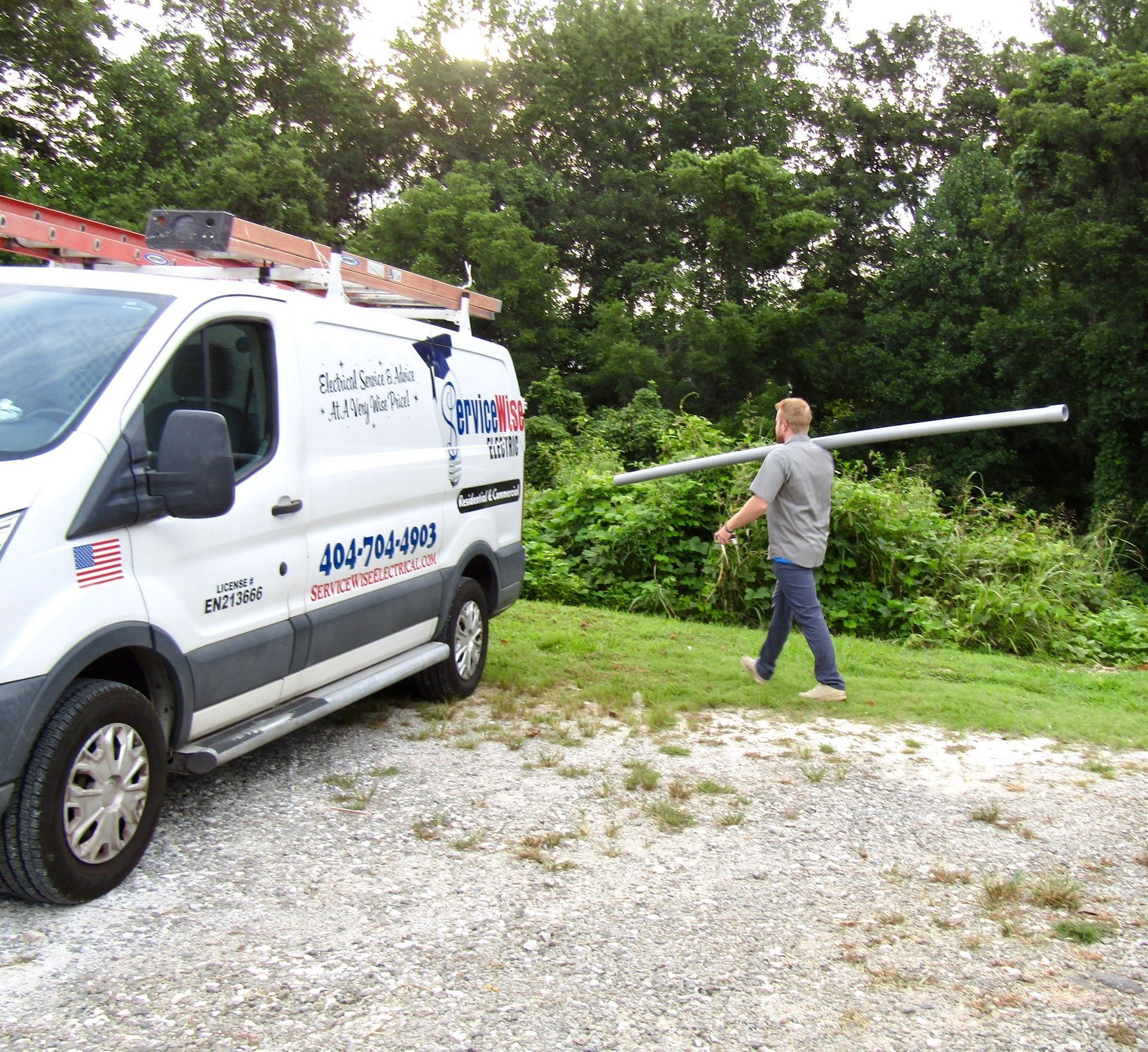 Man carrying a long, white pole near a service van parked on gravel. Green foliage in the background.