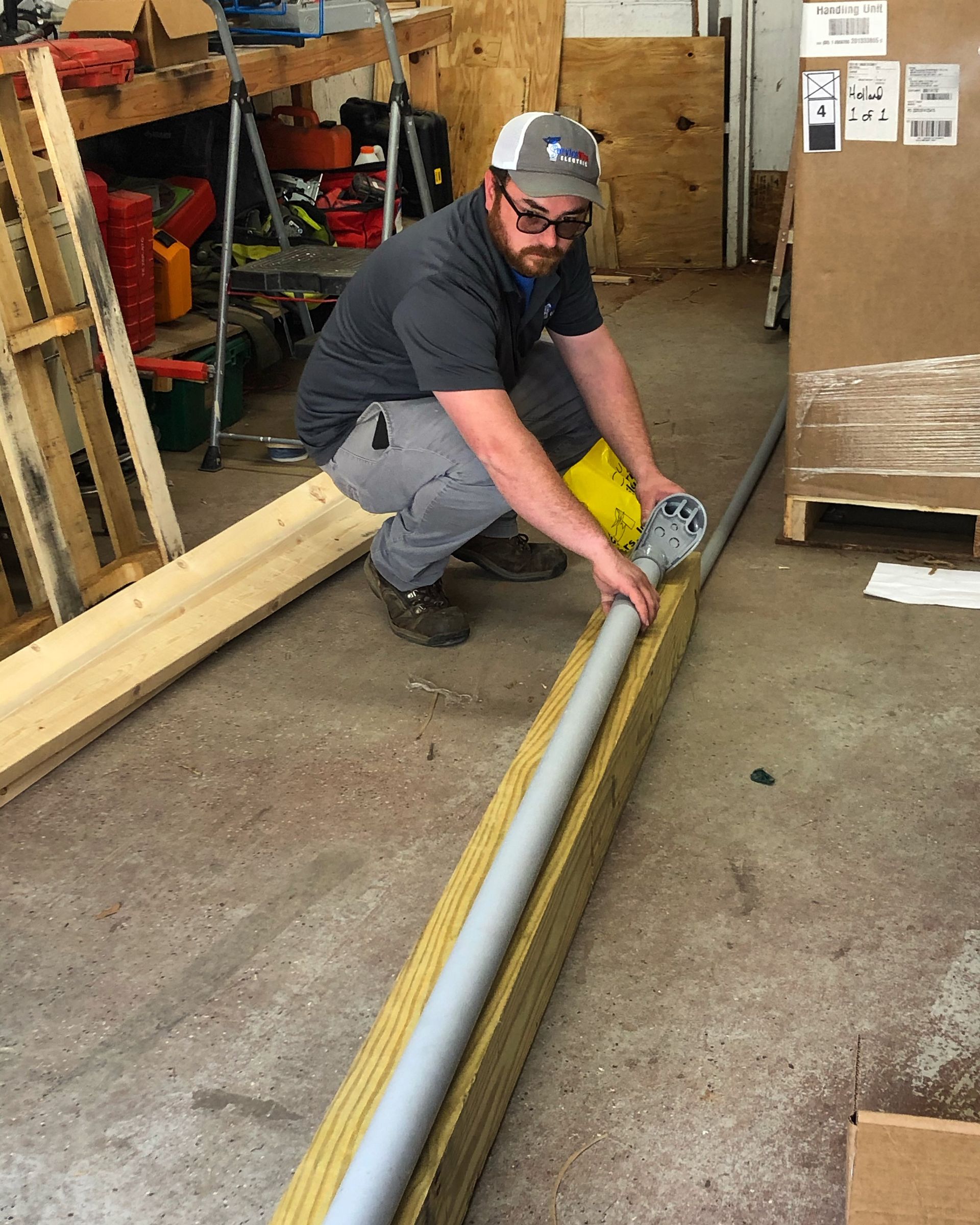 Man in grey shirt working with pipe and wood planks on a concrete floor inside a workshop.