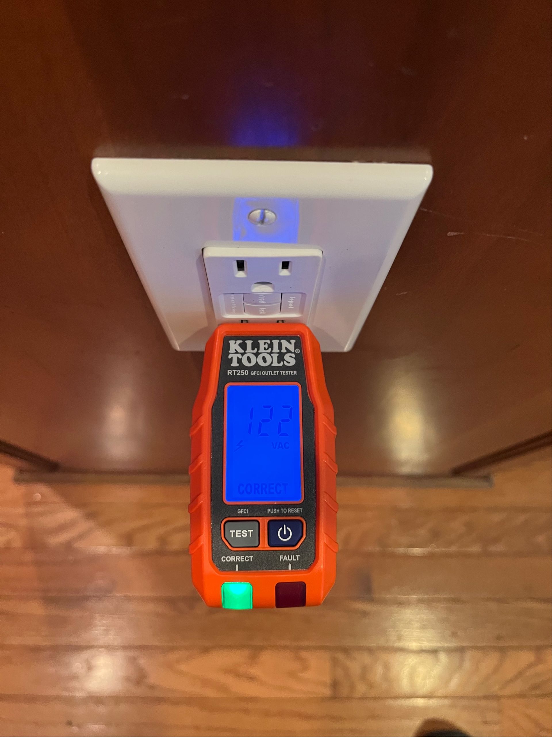 An orange electrical outlet tester plugged into a wall outlet, displaying readings. Green and red indicator lights.