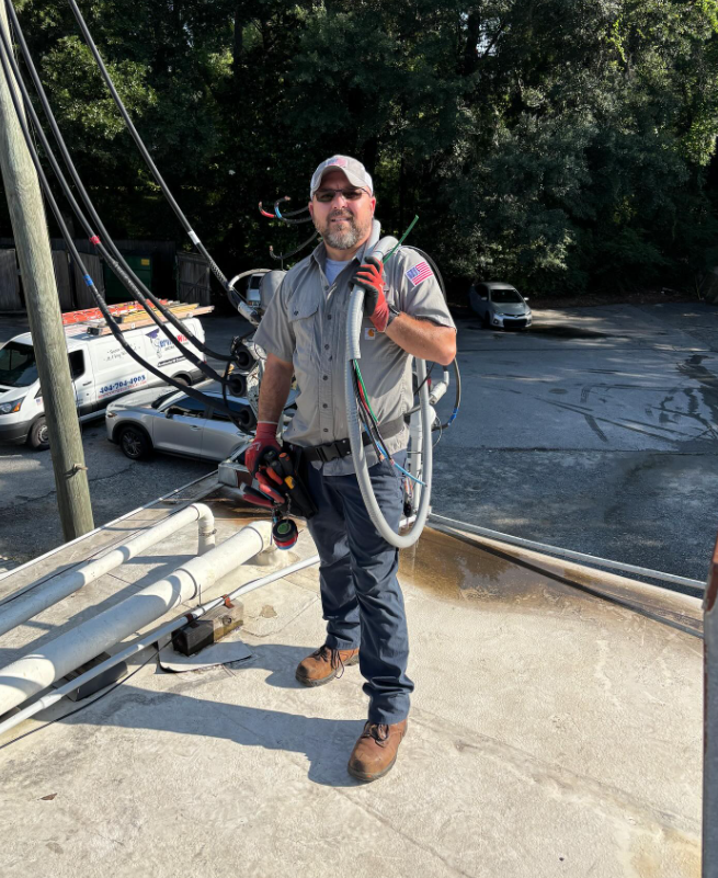Man in work clothes on rooftop, holding a vacuum hose. Utility lines and vehicles in the background.