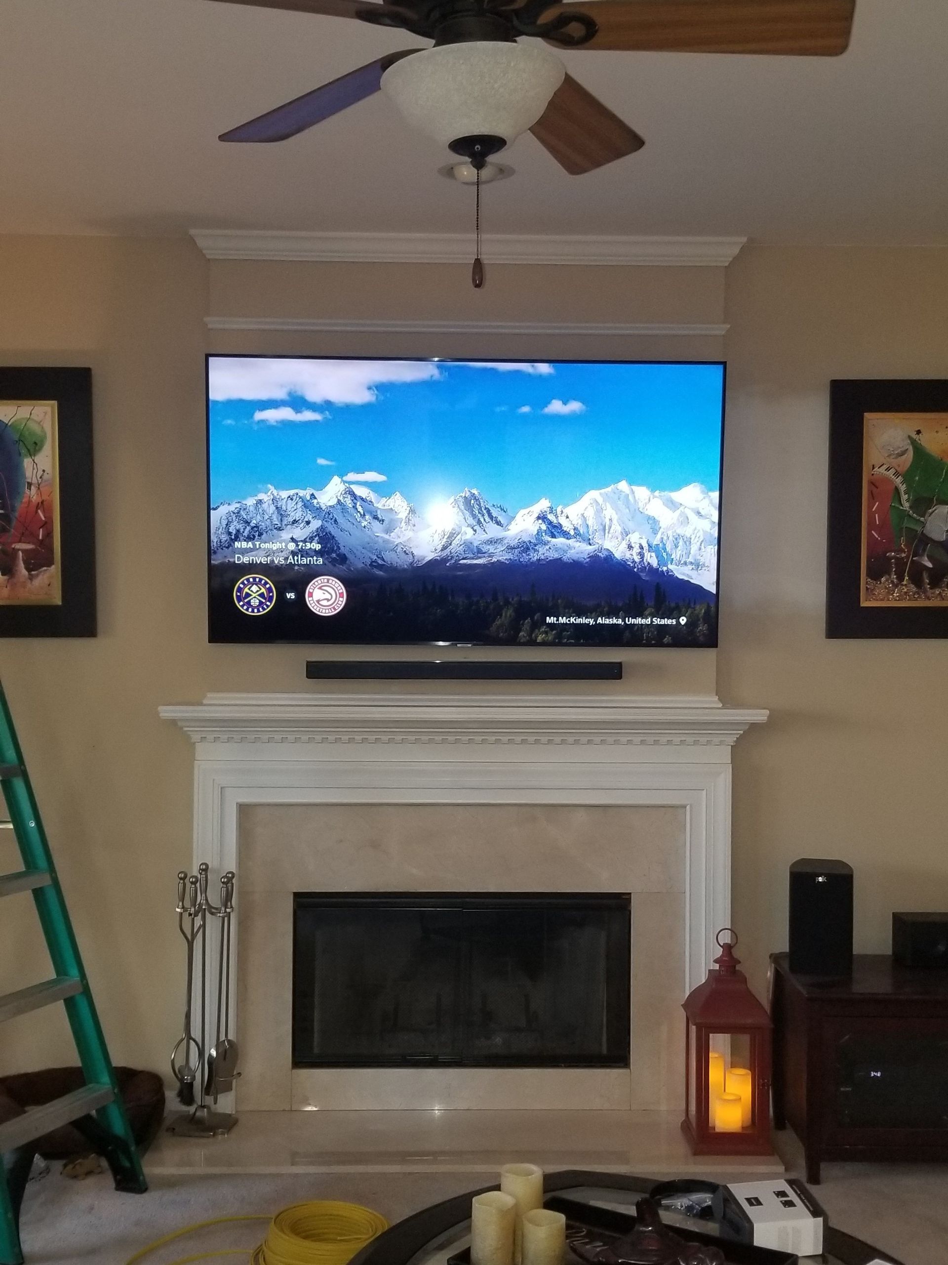TV above fireplace, mountains on screen, flanked by artwork, with a ceiling fan overhead.