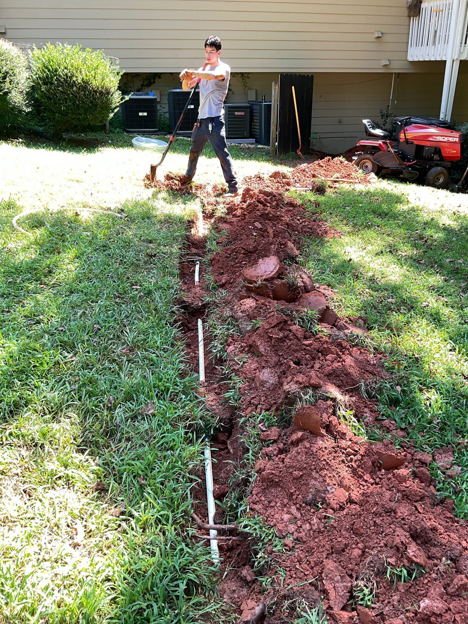 A person digging a trench in a grassy yard, near a building and a riding lawnmower.
