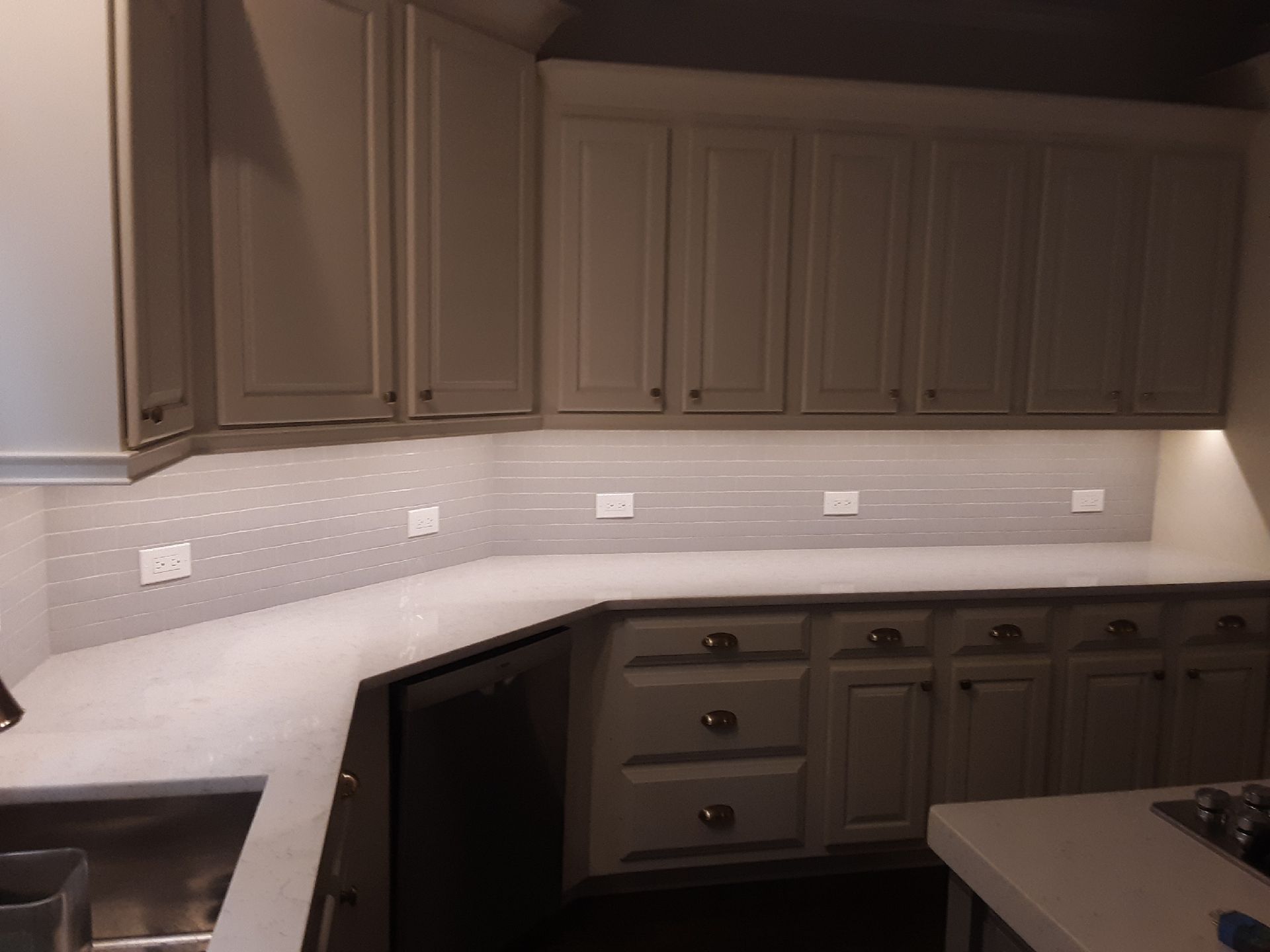 Kitchen cabinets and countertop with under-cabinet lighting. Gray cabinets, light tile backsplash, and white countertop.