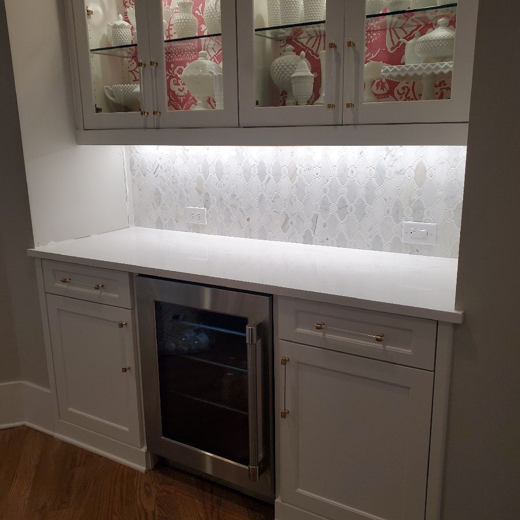 Built-in white cabinets with wine fridge. Glass-front display case above. White countertop with tiled backsplash.