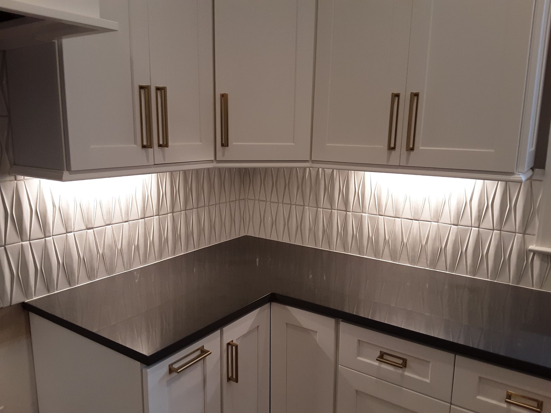 Kitchen corner with white cabinets, dark countertop, textured backsplash, and under-cabinet lighting.