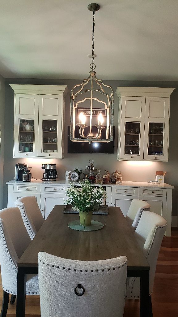 Dining area with a wooden table, upholstered chairs, and a chandelier, with white cabinets and a kitchen counter in the background.