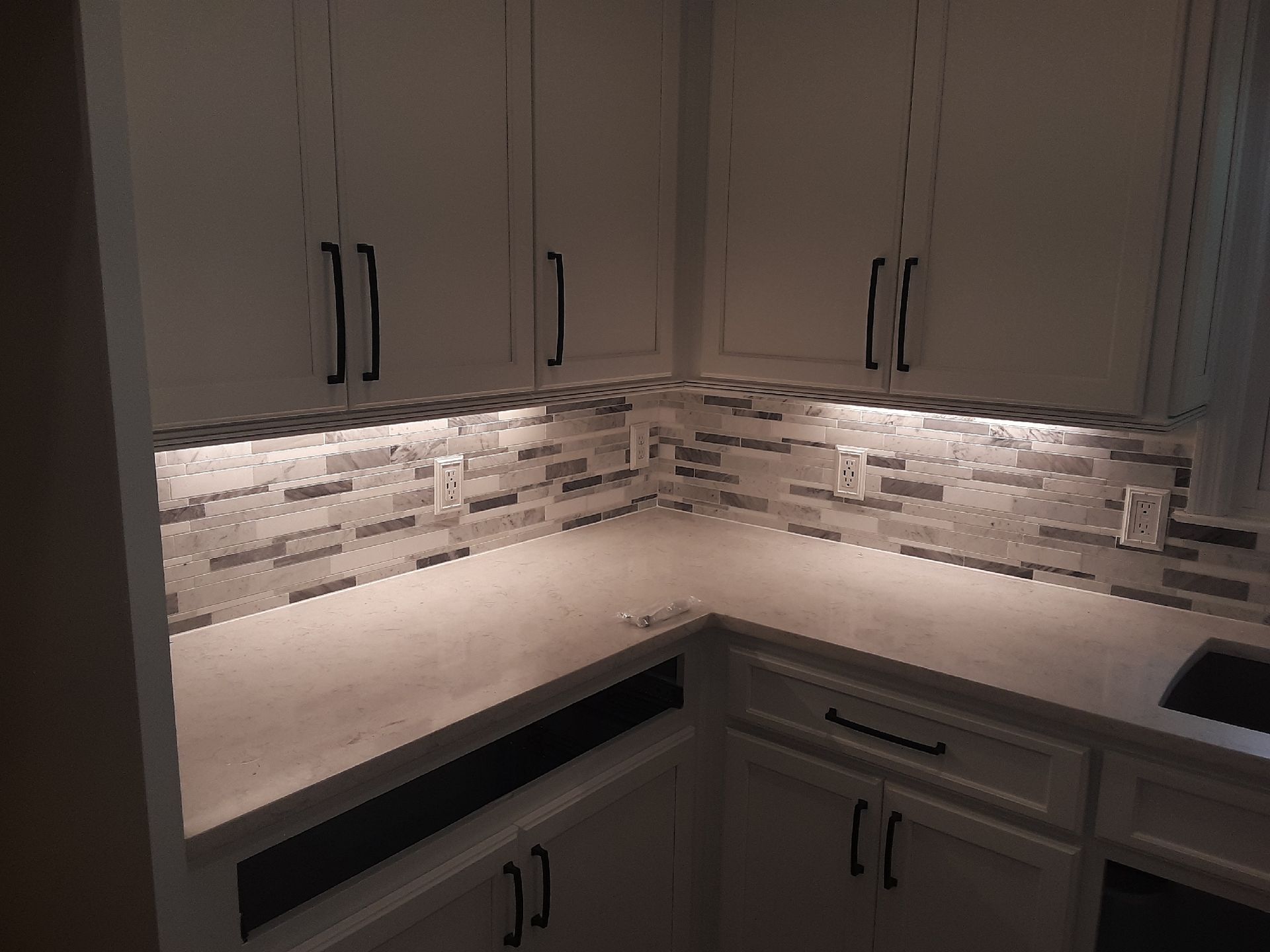 White kitchen cabinets with black handles, gray tiled backsplash, and under-cabinet lighting.