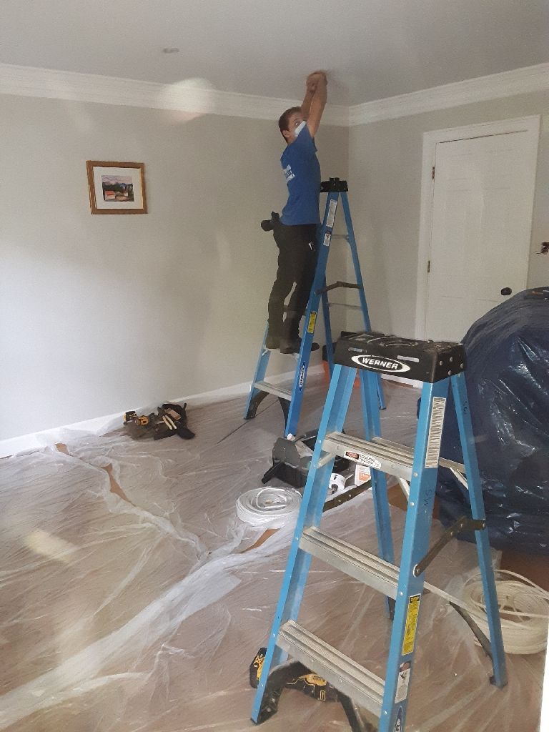 Person on a ladder painting a ceiling in a room with protective coverings on the floor and walls.