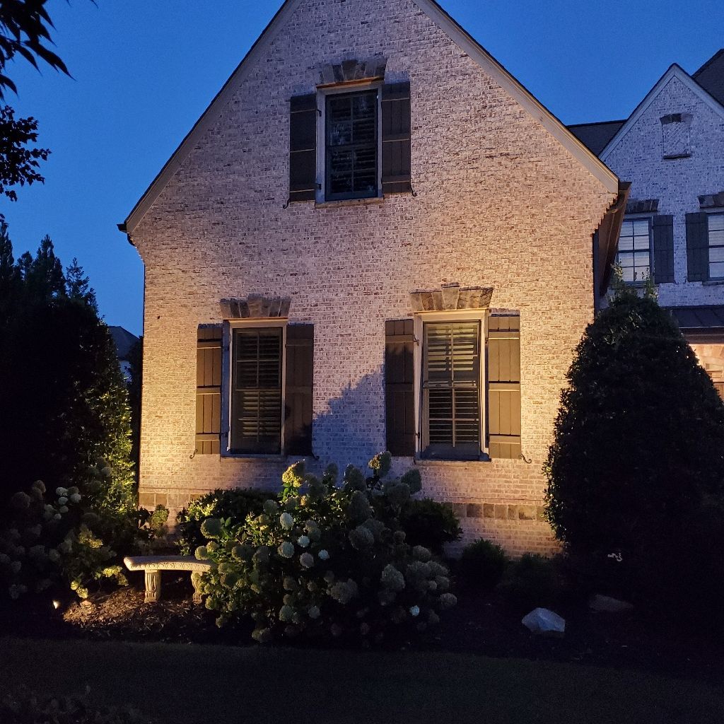 Illuminated brick house with dark shutters, front garden, and blue twilight sky.