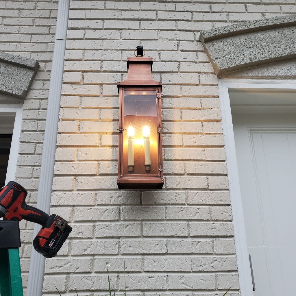 Copper-colored outdoor lantern with two lit candles on a brick wall, next to a white door. A red drill is on a ladder.